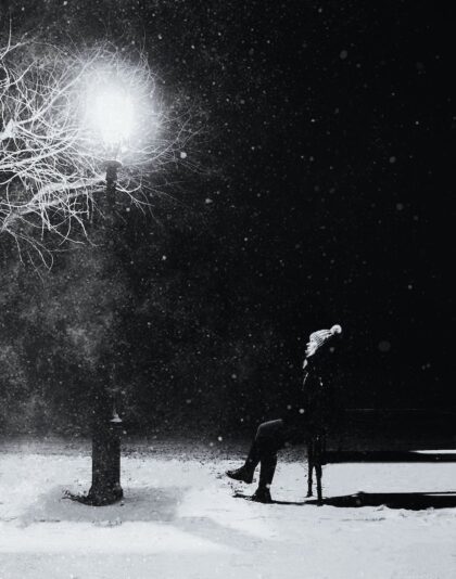 grayscale photography of person with knit pompom cap sits in front turned on light post at night