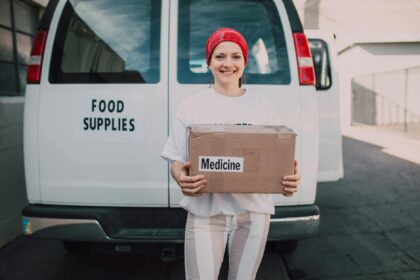 woman carrying a medicine labelled cardboard boxes behind a white van